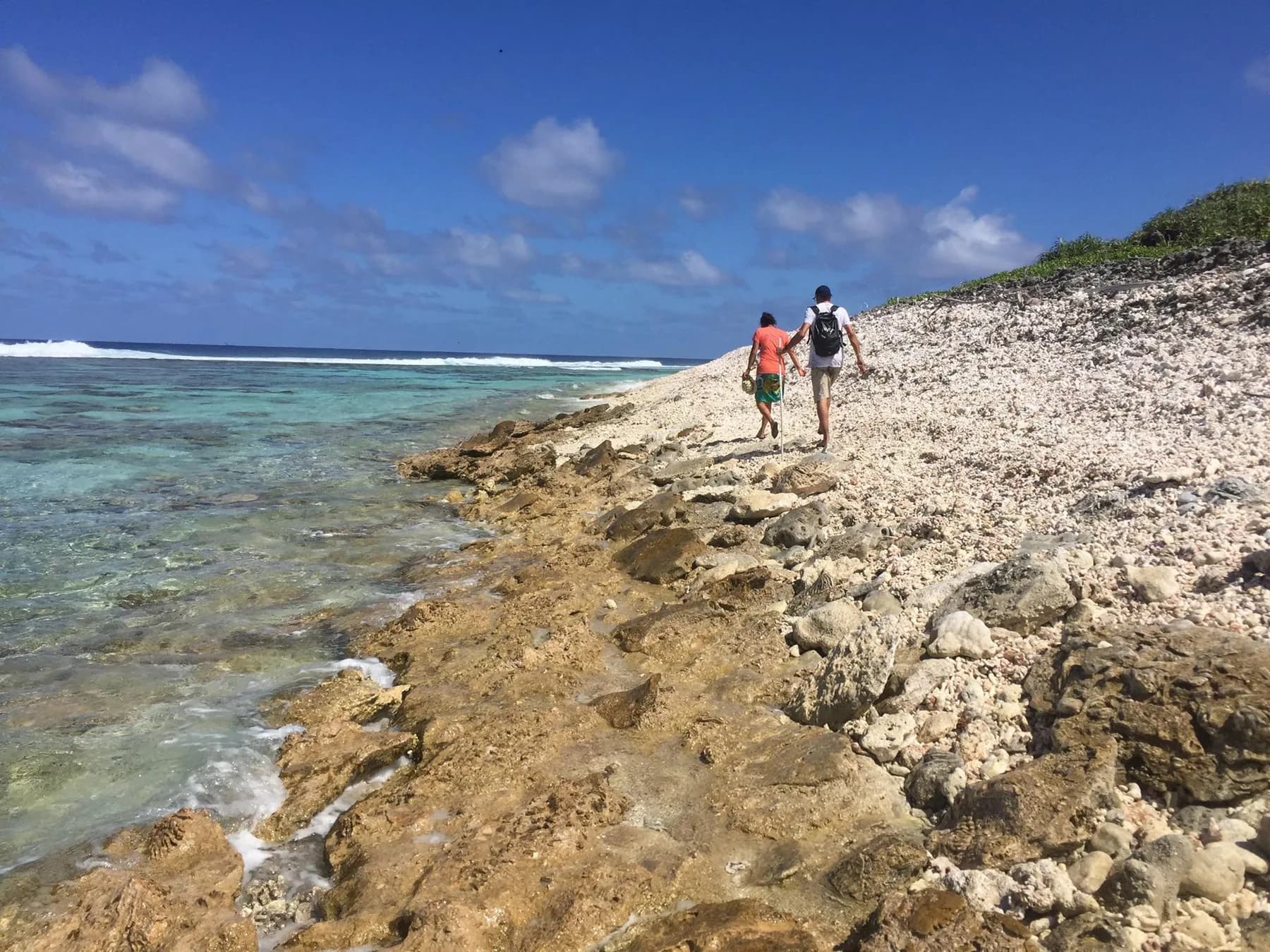 Coral rubble and consolidated reef rock along Tetiaroa’s shore, with two people walking beside the water